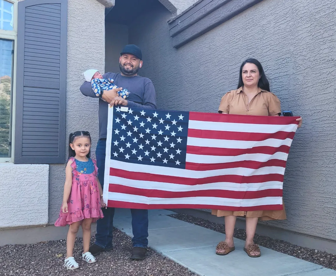 A family of four, including a newborn, holding the American flag in front of their new home.